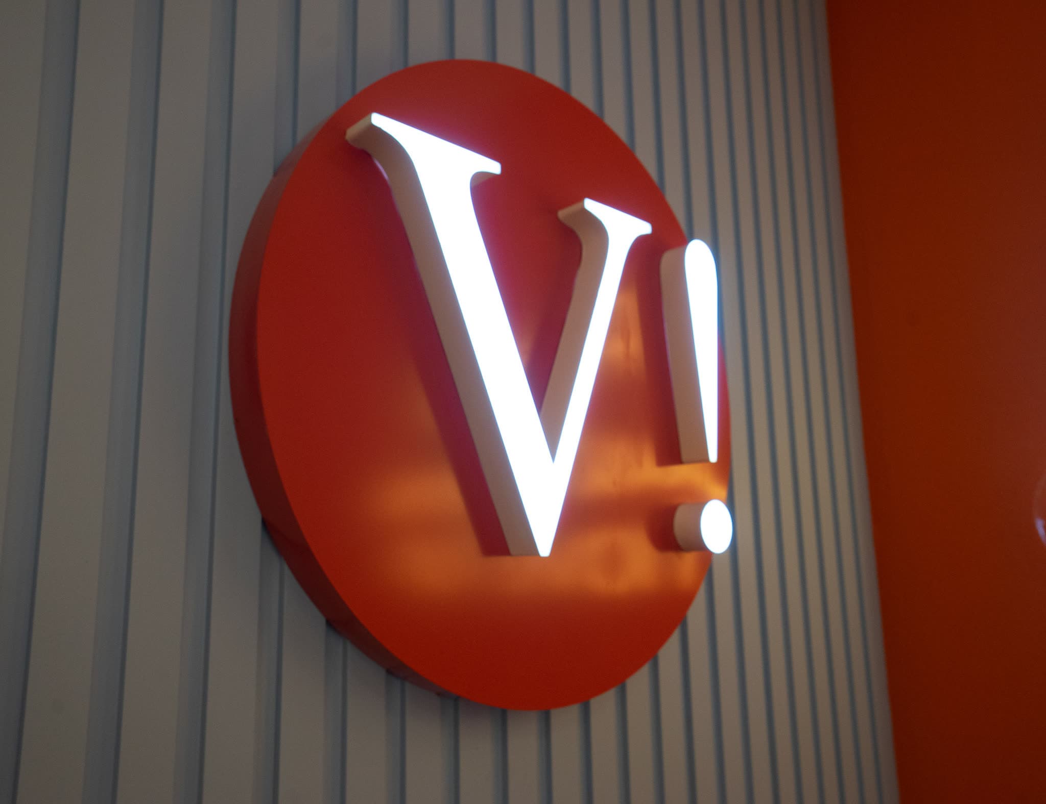 Illuminated built-up letter sign — a red disc with a face-lit V, mounted on a slatted wall