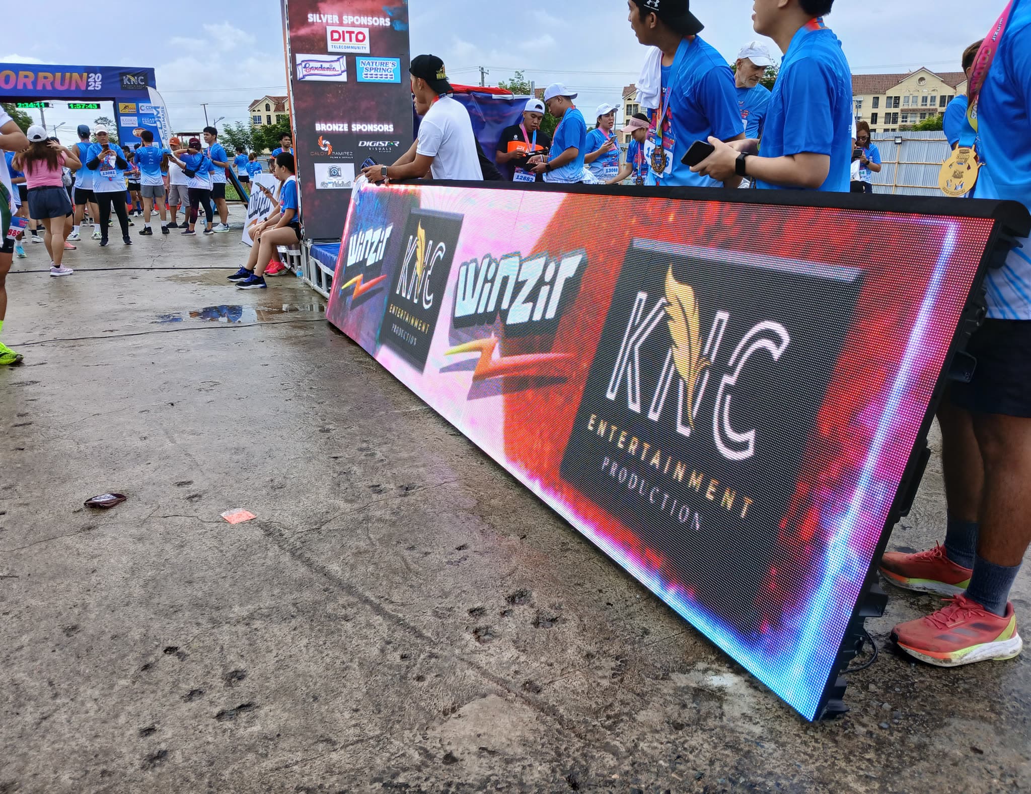 Long LED perimeter banner at a daytime fun-run, with runners gathered behind it
